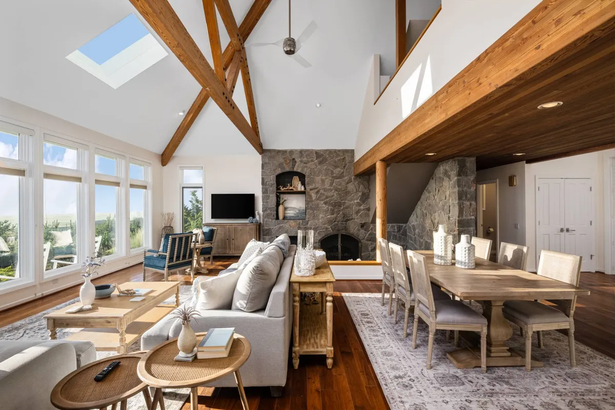 The image showcases a spacious, inviting living and dining area with soaring wooden beams, a dramatic stone fireplace, and expansive windows that frame unobstructed Pacific Ocean views. A large dining table sits ready for family gatherings or holiday feasts, all bathed in warm natural light.