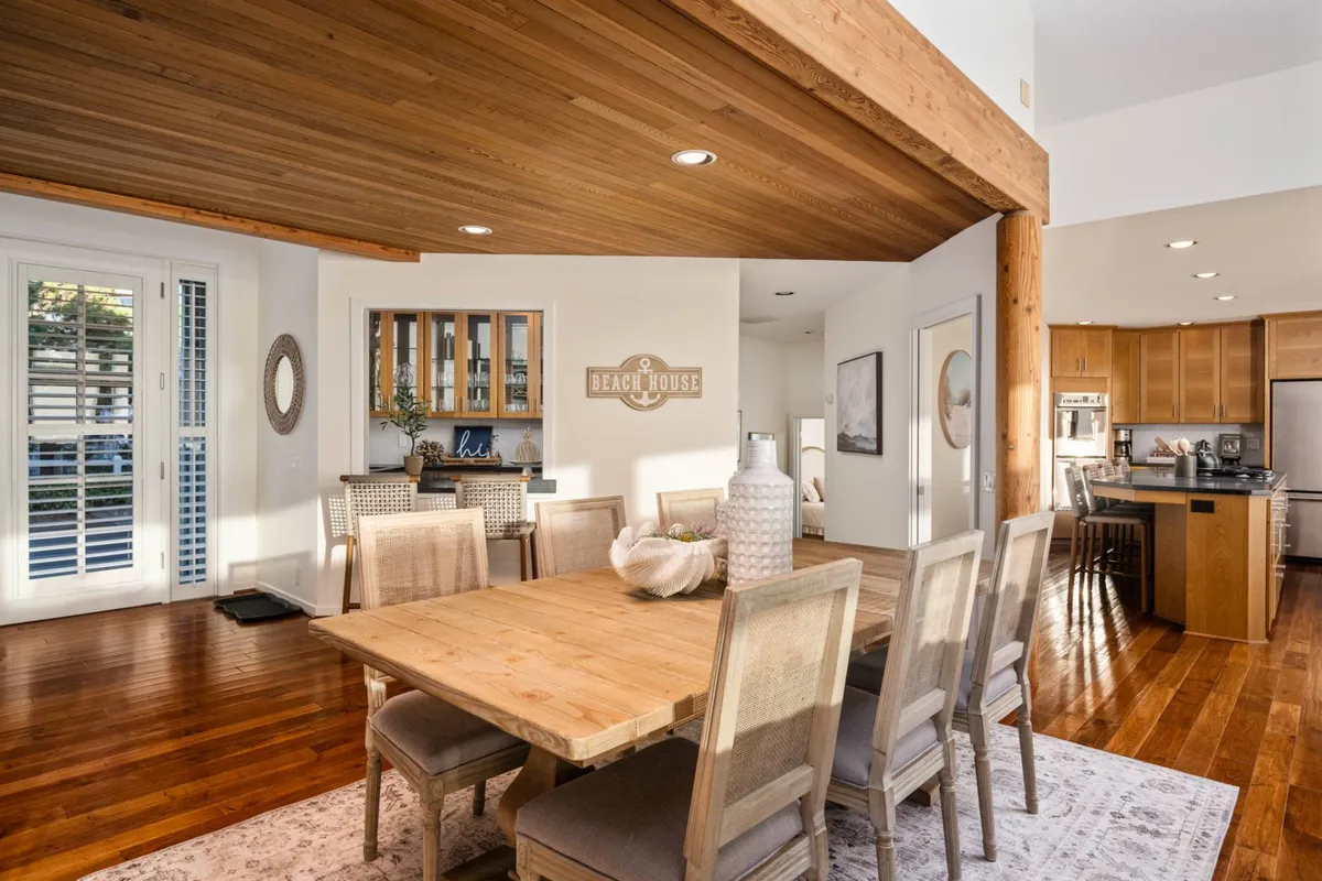A stylish dining area with a wooden table set beneath a vaulted, open-beam ceiling. The space includes a visible kitchen area and a 'beach house' sign, evoking a sense of coastal charm. Large windows allow natural light to flood the room.