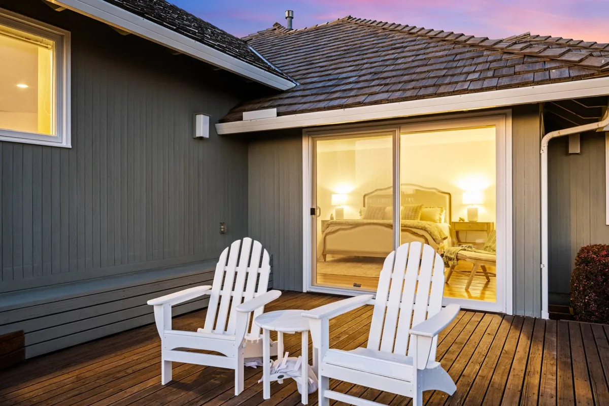 A cozy outdoor patio with white Adirondack chairs, a small round side table, and a view toward the horizon. The warm glow from the illuminated interior and the open sky beyond suggest the perfect spot for enjoying sunsets, sipping evening drinks, or relaxing after a stroll to the ocean.