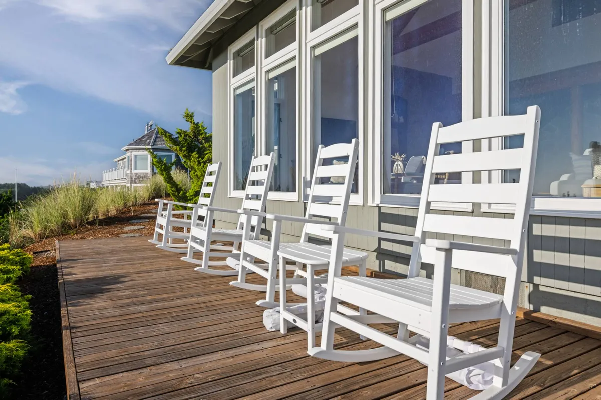 This image showcases a peaceful wooden deck with white rocking chairs positioned to take in the views through large windows. The setting encourages morning coffees or evening chats beneath the stars, seamlessly blending the comfort of home with the allure of the outdoors and regional exploration.
