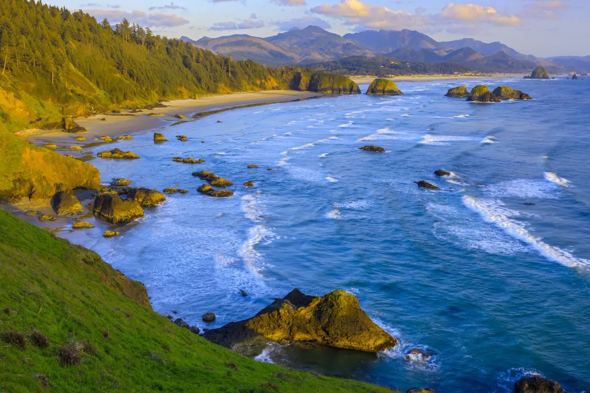 A breathtaking view of a rocky coastline with gentle waves lapping against the sandy beach. Distant mountains and dramatic rock formations frame the scene, conveying a sense of natural adventure and the untamed beauty of the Pacific Northwest shoreline. The beach is inviting for beachcombing and exploration, and the sunset light enhances the tranquil mood.