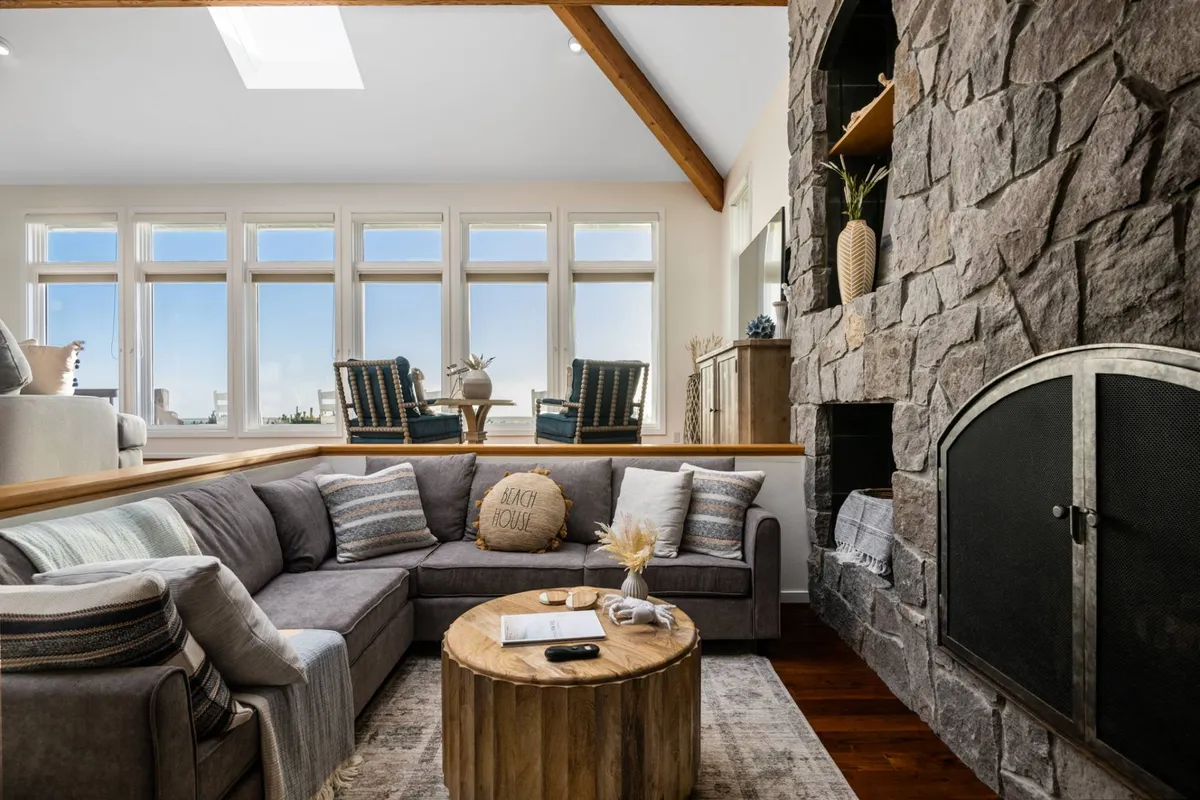 A spacious living area featuring a prominent stone fireplace, a large sectional sofa, a wooden coffee table, and expansive windows that overlook the ocean and fairways. Natural light pours in, creating an inviting environment for relaxation and socializing.