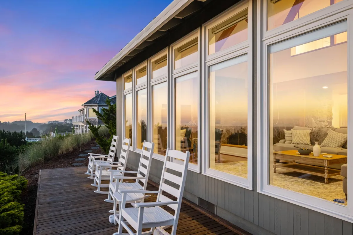 A cozy exterior deck outfitted with rocking chairs, overlooking large windows that reflect a glowing sunset. The interior seating area is visible through the windows, suggesting the retreat’s inviting spaces for gathering and storytelling after a day on the beach.