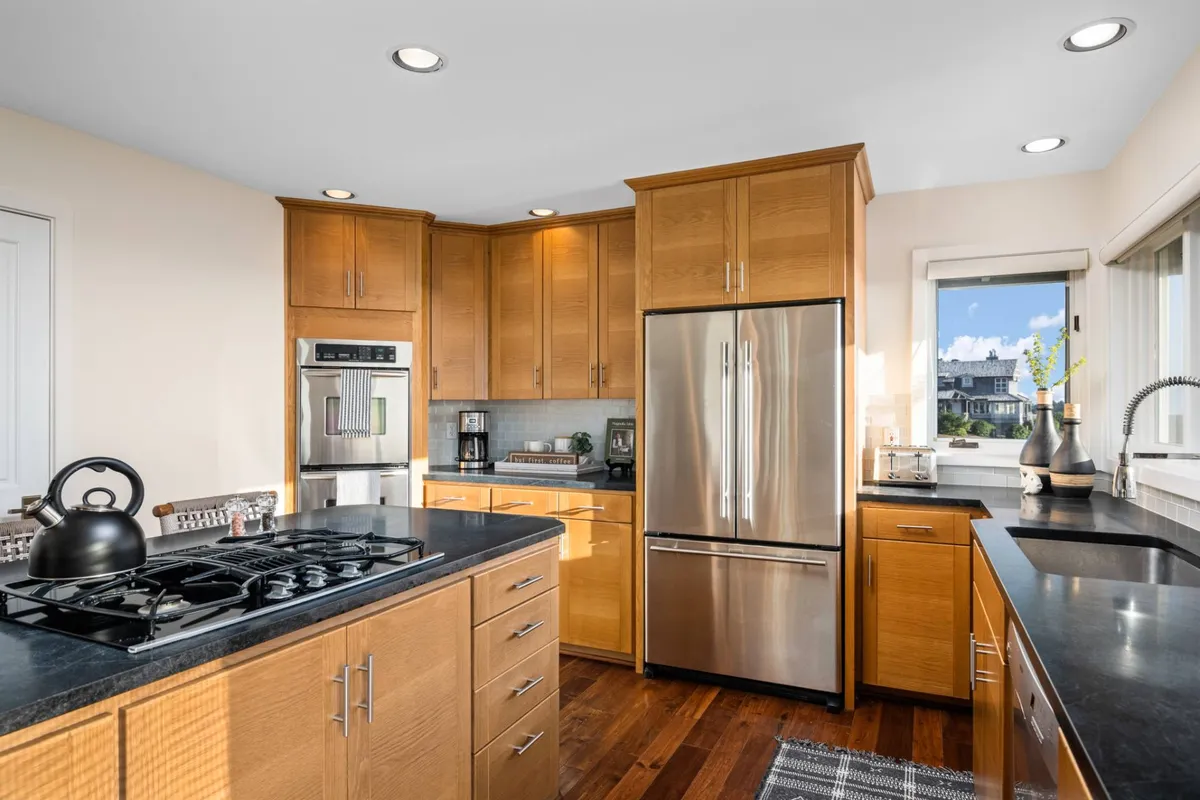 A kitchen scene highlighting a gas stove, stainless steel refrigerator, warm wood cabinetry, and a sleek black countertop, creating an inviting ambiance for cooking and entertaining.