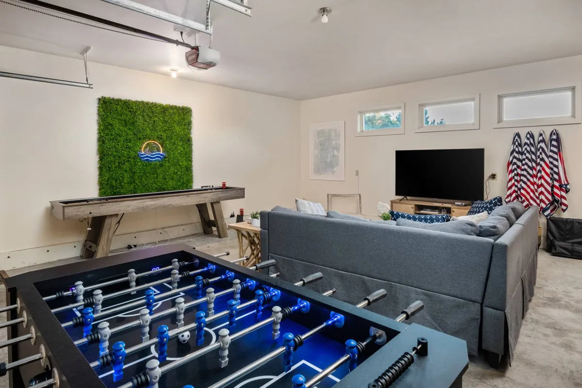 A cozy corner of the game room is shown, featuring a foosball table, sofa, green wall art, and a TV, inviting both players and spectators. The mood is casual and welcoming, perfect for friendly hangouts or movie marathons after games.