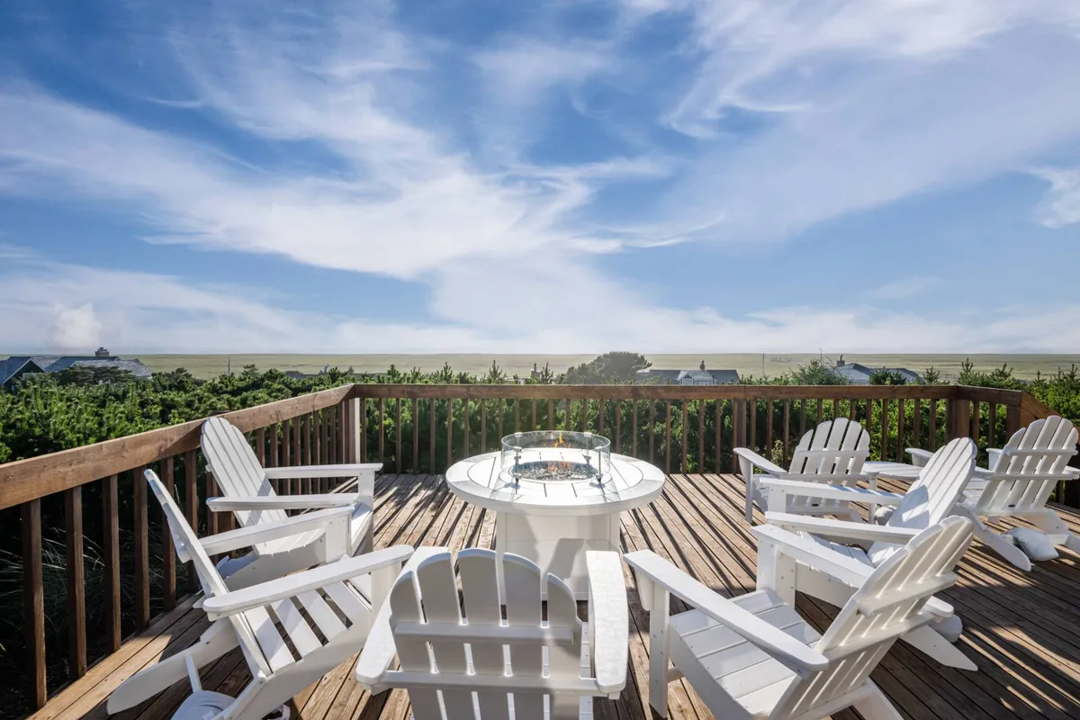 A serene outdoor deck setup featuring white Adirondack chairs and a round table, all facing a breathtaking ocean view. The setting suggests a peaceful spot for unwinding after a day on the course, with coastal panoramas enhancing the relaxing atmosphere.